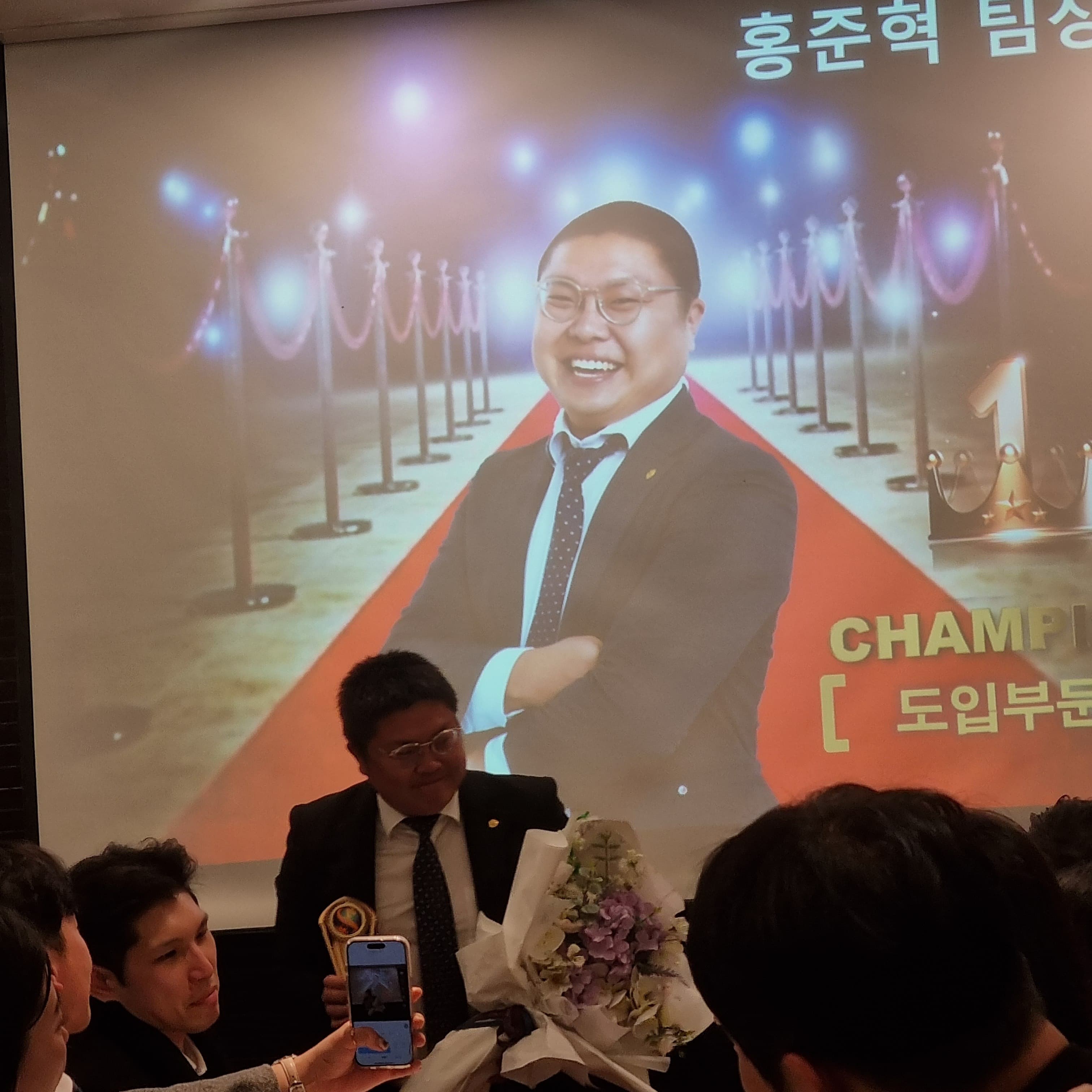 행사 현장에서 홍준혁 소개 화면과 참석자들이 함께 있는 장면